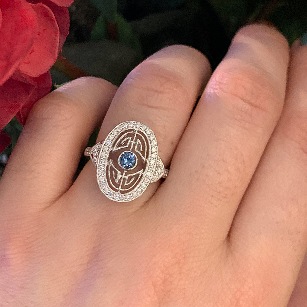 Close-up of an elegant Art Deco style white gold ring featuring a bright blue Yogo sapphire center stone surrounded by   round brilliant-cut diamonds on a person's ring finger.