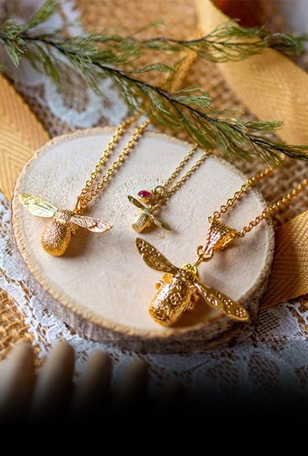 Alex Monroe signature vermeil bee necklaces on a wooden surface with greenery