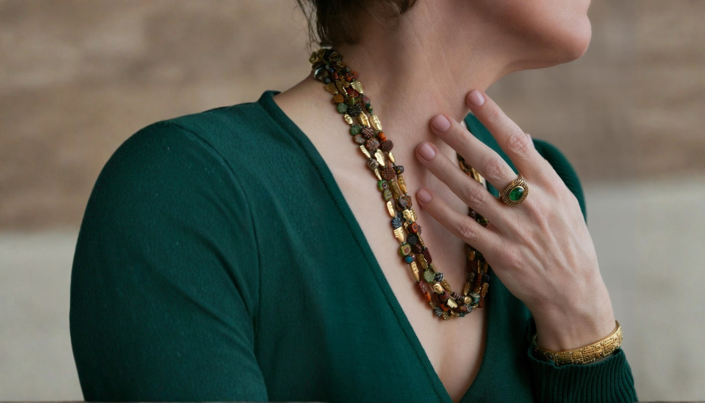 A close-up lifestyle shot of a woman wearing a multi-strand chunky necklace with gold and brown beads, a green ring, and a gold bracelet, wearing a dark green top.