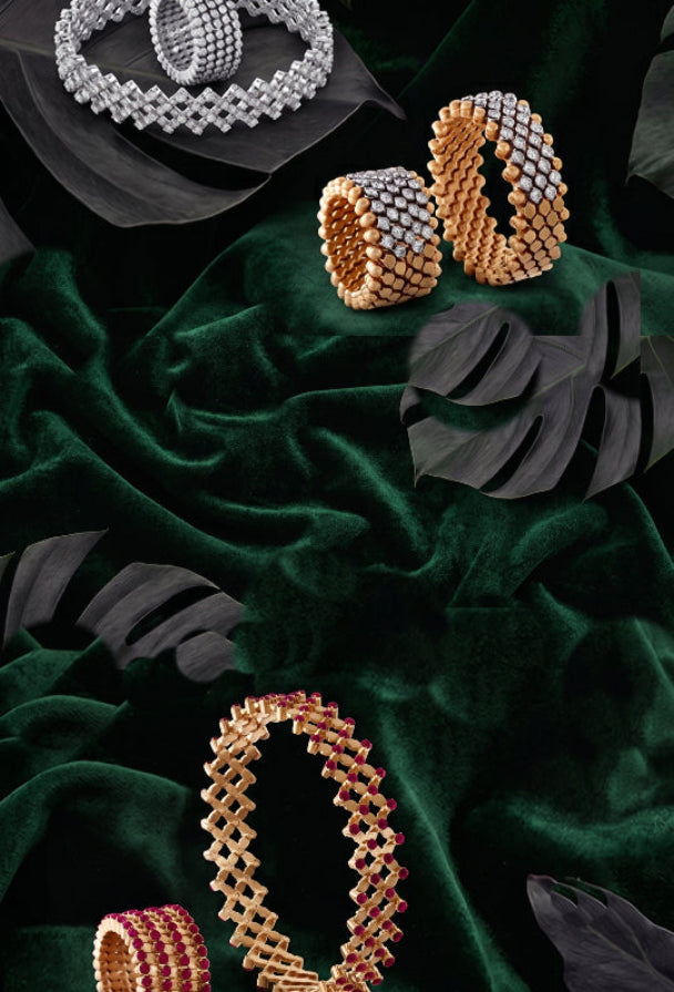 A luxury jewelry flat-lay featuring multiple woven mesh bands in gold, silver, and pink-gold tones, embellished with sparkling clear stones, arranged on a draped dark green velvet fabric with black monstera leaves.