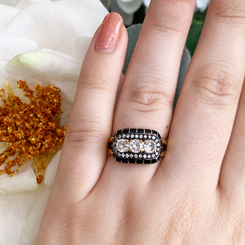 A hand wearing a 18k gold ring featuring 3 brilliant-cut round center diamonds surrounded by a halo of smaller round diamonds and wrapped in black enamel.